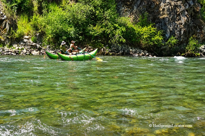 Double J&D Historic Hot Spring Ranch in Idaho: The picture "green inflatable raft on clear river" depicts two people riding a bright green inflatable raft down a clear, fast-flowing river, bordered by steep rocky cliffs and dense greenery.