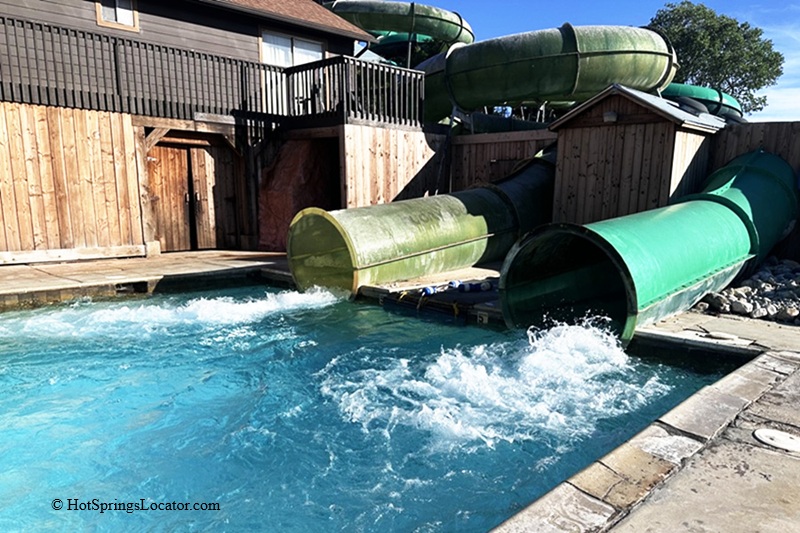 Crystal Hot Springs in Utah - Water Slides at Crystal Hot Springs Crystal Hot Springs in Utah: The picture "Water Slides at Crystal Hot Springs" features two large, green enclosed waterslides emptying into a square blue pool, surrounded by wooden fencing and shaded by trees.