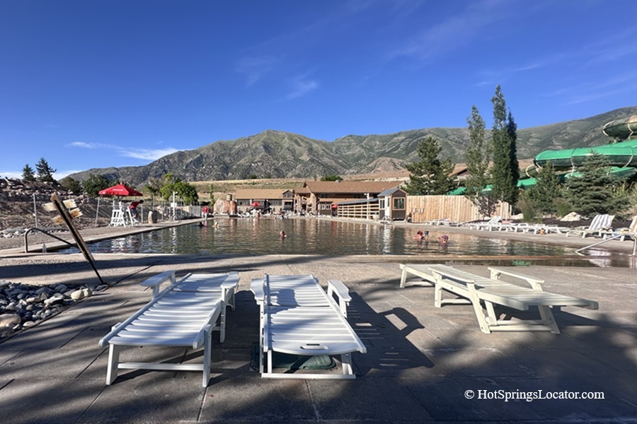 Crystal Hot Springs in Utah - Crystal Hot Springs Main Pool Crystal Hot Springs in Utah: The picture "Crystal Hot Springs Main Pool" shows a large swimming pool framed by white lounge chairs on a sunny concrete deck, with mountains rising in the background and several people enjoying the water near the far end.