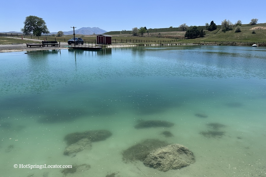 Belmont Hot Springs (Formerly Udy Hot Springs): Utah's Geothermal Resort in northern Utah - Belmont Hot Springs Main Pool Belmont Hot Springs Main Pool shows a large, turquoise-colored geothermal pond with crystal clear water and submerged rocks, surrounded by grassy banks, a dock, a small red building, and hills in the distance.