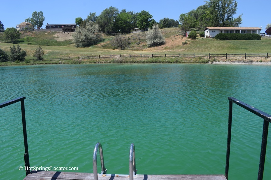 Belmont Hot Springs (Formerly Udy Hot Springs): Utah's Geothermal Resort in northern Utah - Poolside View at Belmont Hot Springs Poolside View at Belmont Hot Springs displays the pond from the perspective of a metal pool ladder attached to a dock, looking across the emerald surface toward landscaped grounds and a row of houses beyond a fence.