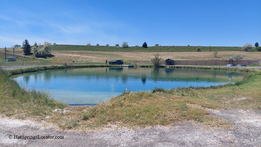 Belmont Hot Springs (Formerly Udy Hot Springs): Utah's Geothermal Resort in northern Utah - Belmont Hot Springs Pond Overview Belmont Hot Springs Pond Overview presents a wide view of the green-blue pond bordered by grass, with a few people and a car near the opposite shore, surrounded by gently rolling hills on a clear day.