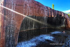 Crystal Hot Springs in Utah - Waterfall at Crystal Hot Springs Crystal Hot Springs in Utah: The picture "Waterfall at Crystal Hot Springs" captures a close-up of the hot spring’s artificial red rock wall with steaming water cascading down into the pool below, where a swimmer enjoys the falls.