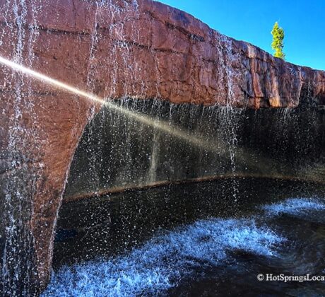 Crystal Hot Springs in Utah - Waterfall at Crystal Hot Springs Crystal Hot Springs in Utah: The picture "Waterfall at Crystal Hot Springs" captures a close-up of the hot spring’s artificial red rock wall with steaming water cascading down into the pool below, where a swimmer enjoys the falls.