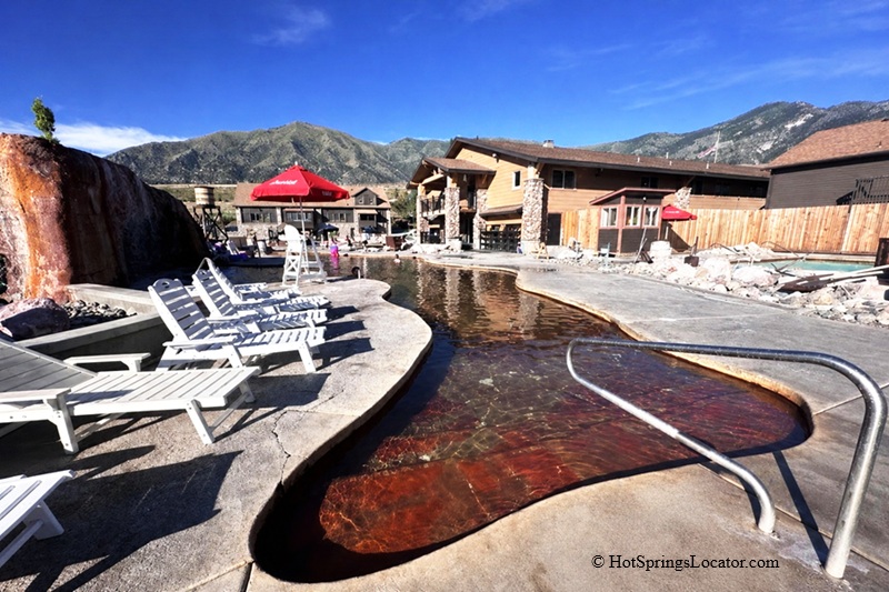 Crystal Hot Springs in Utah - Crystal Hot Springs Hot Pool Crystal Hot Springs in Utah: The picture "Crystal Hot Springs Hot Pool" shows a smaller, winding mineral pool with rusty orange water, rimmed by white lounge chairs, and flanked by a faux red rock waterfall and the main bathhouse.