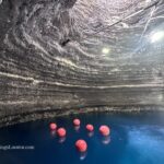 Homestead Crater Hot Springs in Utah: The picture "Homestead Crater Interior" displays the inside of the natural dome-shaped crater with blue mineral water and several floating red buoys, illuminated by overhead lights attached to the rock walls.