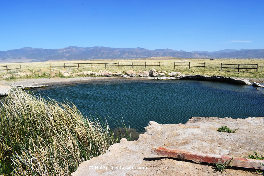 Meadow Hot Springs in Utah: The picture "Meadow Hot Springs Pool View" shows a large, clear blue natural hot spring pool edged with rocky ledges and grass, set in an open meadow with wooden fences and distant mountains under a clear blue sky.