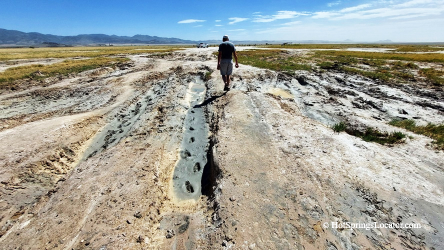 Meadow Hot Springs in Utah: The picture "Meadow Hot Springs Walkway" features a person walking along a muddy, rutted path across an open field toward the hot springs, with meadows and mountains stretching out in the background.