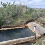 Baker Hot Springs in Utah: The picture "Baker Hot Springs Pool by Creek" shows a rectangular concrete soaking pool next to a small stream, bordered by thick grass and shrubs, under an overcast sky.
