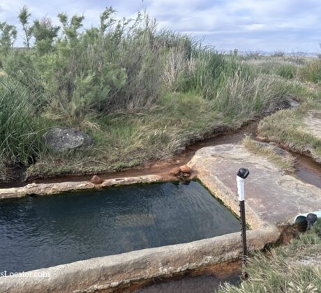Baker Hot Springs in Utah: The picture "Baker Hot Springs Pool by Creek" shows a rectangular concrete soaking pool next to a small stream, bordered by thick grass and shrubs, under an overcast sky.