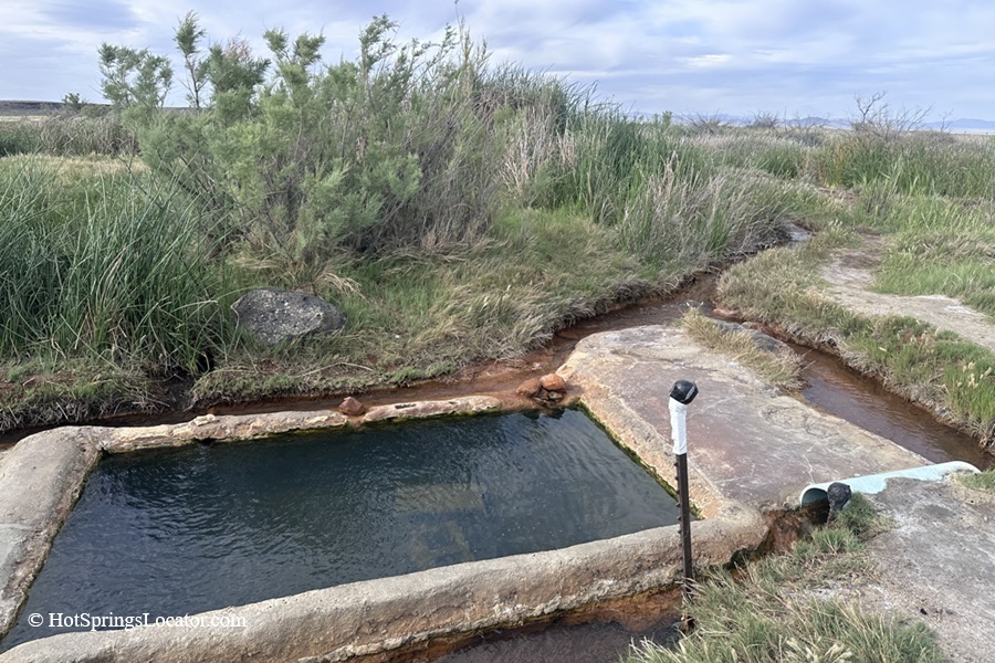 Baker Hot Springs in Utah: The picture "Baker Hot Springs Pool by Creek" shows a rectangular concrete soaking pool next to a small stream, bordered by thick grass and shrubs, under an overcast sky.