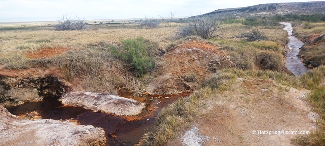 Baker Hot Springs in Utah – Baker Hot Springs Source Creek Baker Hot Springs in Utah: The picture "Baker Hot Springs Source Creek" captures the reddish, mineral-rich source creek winding through rough, grassy terrain and barren patches, with hills visible in the background.