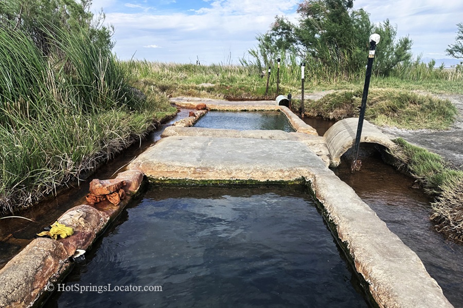 Baker Hot Springs in Utah: The picture "Baker Hot Springs Pools Close-Up" shows rectangular concrete soaking pools filled with geothermal water, surrounded by grassy vegetation and small lamp posts under a partly cloudy sky.