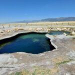 Meadow Hot Springs in Utah: The picture "Meadow Hot Springs Main Pool" displays the main thermal pool with turquoise water bordered by pale, rocky edges, situated in a grassy meadow with wooden fencing and mountains beyond.