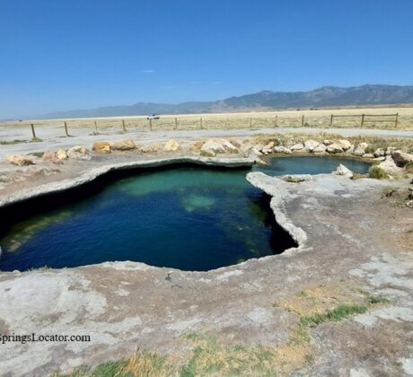 Meadow Hot Springs in Utah: The picture "Meadow Hot Springs Main Pool" displays the main thermal pool with turquoise water bordered by pale, rocky edges, situated in a grassy meadow with wooden fencing and mountains beyond.