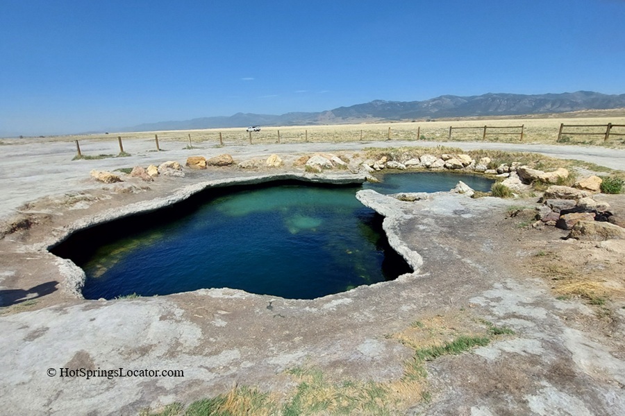 Meadow Hot Springs in Utah: The picture "Meadow Hot Springs Main Pool" displays the main thermal pool with turquoise water bordered by pale, rocky edges, situated in a grassy meadow with wooden fencing and mountains beyond.