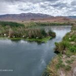 Horseshoe Springs in Utah – Horseshoe Springs Marsh View Horseshoe Springs in Utah – The picture "Horseshoe Springs Marsh View" shows a calm spring-fed pond surrounded by green reeds and grasses, with distant dry hills and rugged mountains under a cloudy sky.