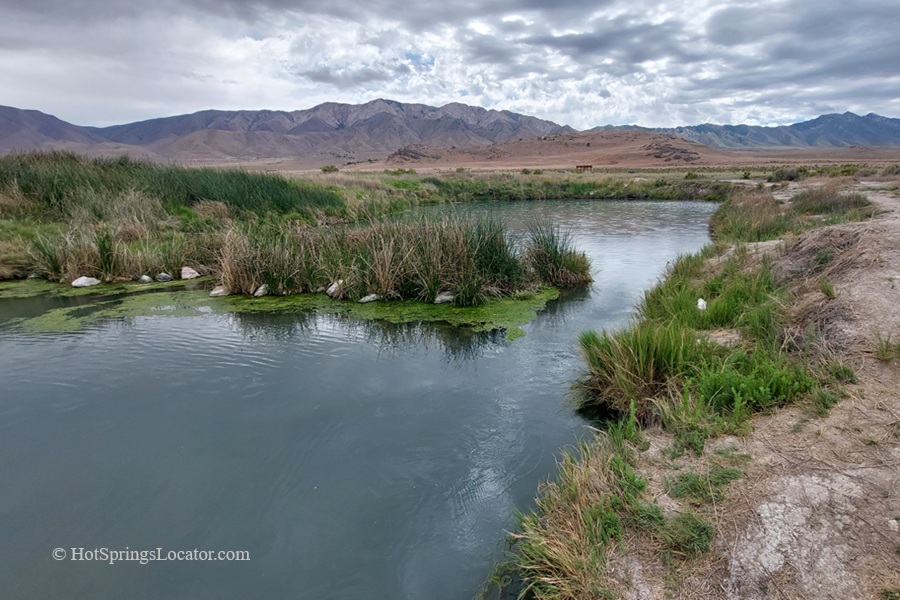 Horseshoe Springs in Utah – Horseshoe Springs Marsh ViewHorseshoe Springs in Utah – The picture "Horseshoe Springs Marsh View" shows a calm spring-fed pond surrounded by green reeds and grasses, with distant dry hills and rugged mountains under a cloudy sky.
