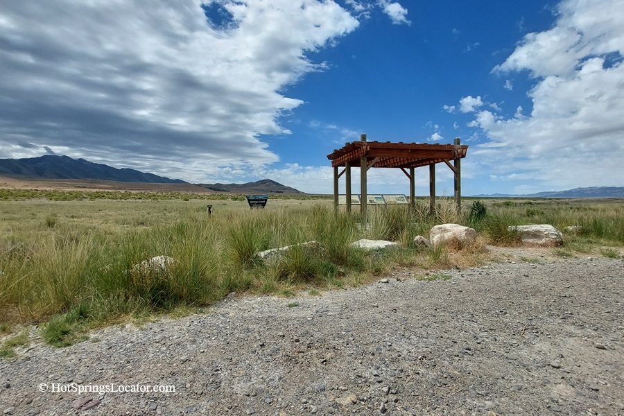 Horseshoe Springs in Utah – Horseshoe Springs Picnic Shelter Horseshoe Springs in Utah – The picture "Horseshoe Springs Picnic Shelter" displays a small wooden picnic shelter standing next to informational signs in a wide, grassy field, with scattered rocks, gravel, and mountains visible beneath a partly cloudy blue sky.