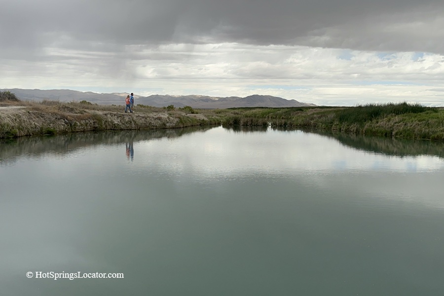 Horseshoe Springs in Utah – Horseshoe Springs Pond ReflectionHorseshoe Springs in Utah – The picture "Horseshoe Springs Pond Reflection" features the glassy surface of a wide pond reflecting cloudy skies, with two people walking along the natural, grassy bank in the distance.