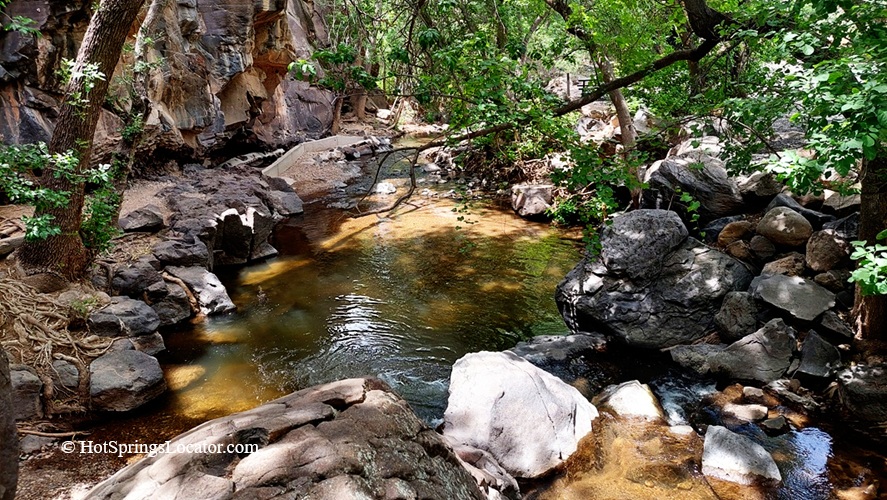 Veyo Pool Resort and Hot Spring, Utah – Veyo Creek and Rock Ledge Veyo Pool Resort and Hot Spring, Utah – The picture "Veyo Creek and Rock Ledge" shows a shallow, gently flowing creek winding through large boulders and trees, with sunlight dappling the water and a rocky cliffside in the background.