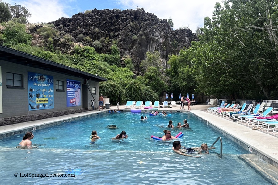 Veyo Pool Resort and Hot Spring, Utah – Veyo Pool Rocky Hillside and Swimmers Veyo Pool Resort and Hot Spring, Utah – The picture "Veyo Pool Rocky Hillside and Swimmers" features people enjoying a large outdoor pool with pool noodles and inflatables, bordered by a rocky volcanic hillside and lined with turquoise lounge chairs and umbrellas.
