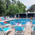 Veyo Pool Resort and Hot Spring, Utah – Veyo Pool Loungers and Swimming Area Veyo Pool Resort and Hot Spring, Utah – The picture "Veyo Pool Loungers and Swimming Area" displays a lively outdoor swimming pool with adults and children swimming or lounging on inflatable floats, surrounded by rows of blue and white lounge chairs under a partly cloudy sky and shaded by tall trees.