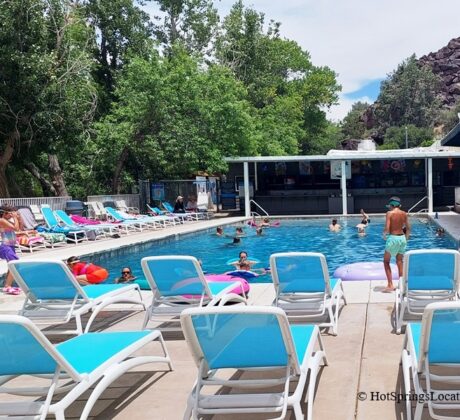 Veyo Pool Resort and Hot Spring, Utah – Veyo Pool Loungers and Swimming Area Veyo Pool Resort and Hot Spring, Utah – The picture "Veyo Pool Loungers and Swimming Area" displays a lively outdoor swimming pool with adults and children swimming or lounging on inflatable floats, surrounded by rows of blue and white lounge chairs under a partly cloudy sky and shaded by tall trees.