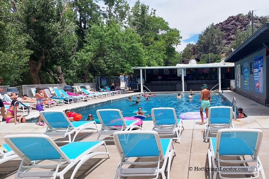 Veyo Pool Resort and Hot Spring, Utah – Veyo Pool Loungers and Swimming Area Veyo Pool Resort and Hot Spring, Utah – The picture "Veyo Pool Loungers and Swimming Area" displays a lively outdoor swimming pool with adults and children swimming or lounging on inflatable floats, surrounded by rows of blue and white lounge chairs under a partly cloudy sky and shaded by tall trees.